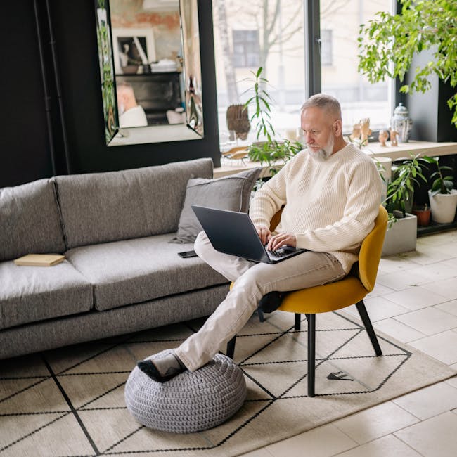 Elderly man in sweater using laptop at home in modern living room, daytime.